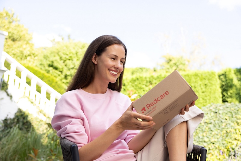 Una donna sorridente è seduta in giardino e tiene in mano un pacco di Shop Apotheke.